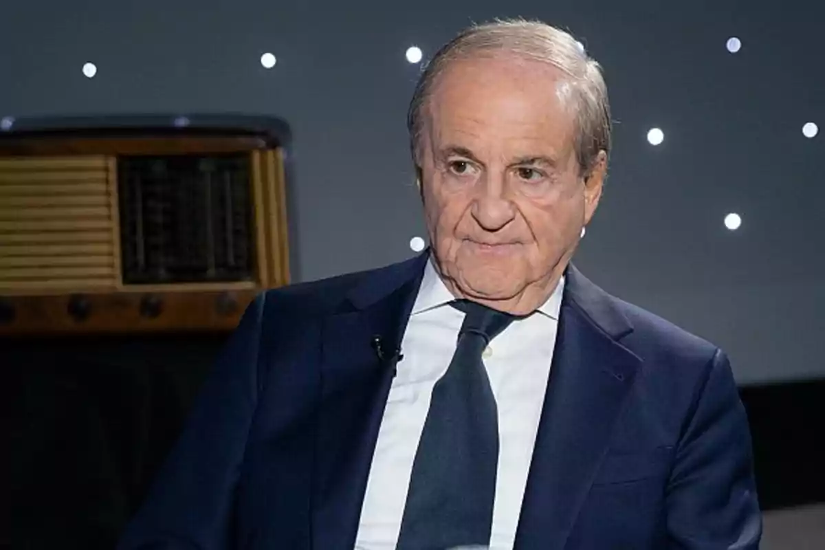 An older man in a dark suit and tie is sitting in front of a dark background with small lights. An older man in a dark suit and tie is sitting in front of a dark background with small lights.