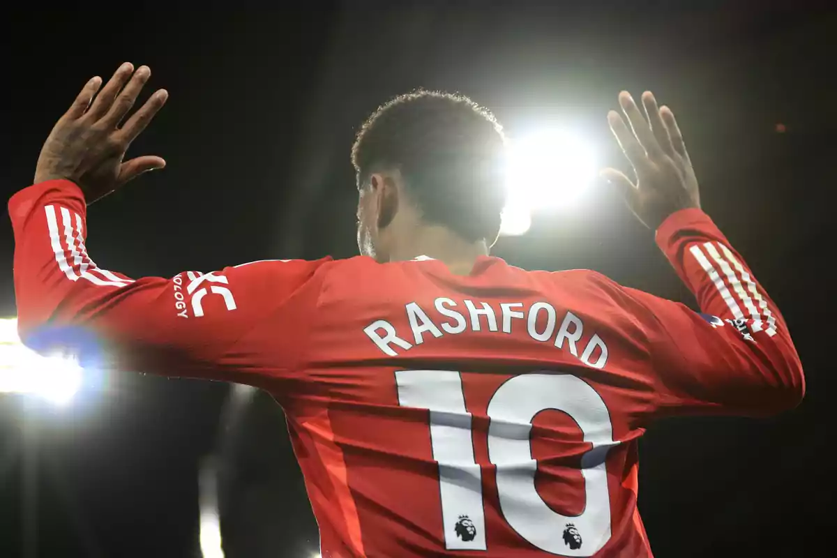 Football player in red jersey number 10 raising his hands in front of bright lights.