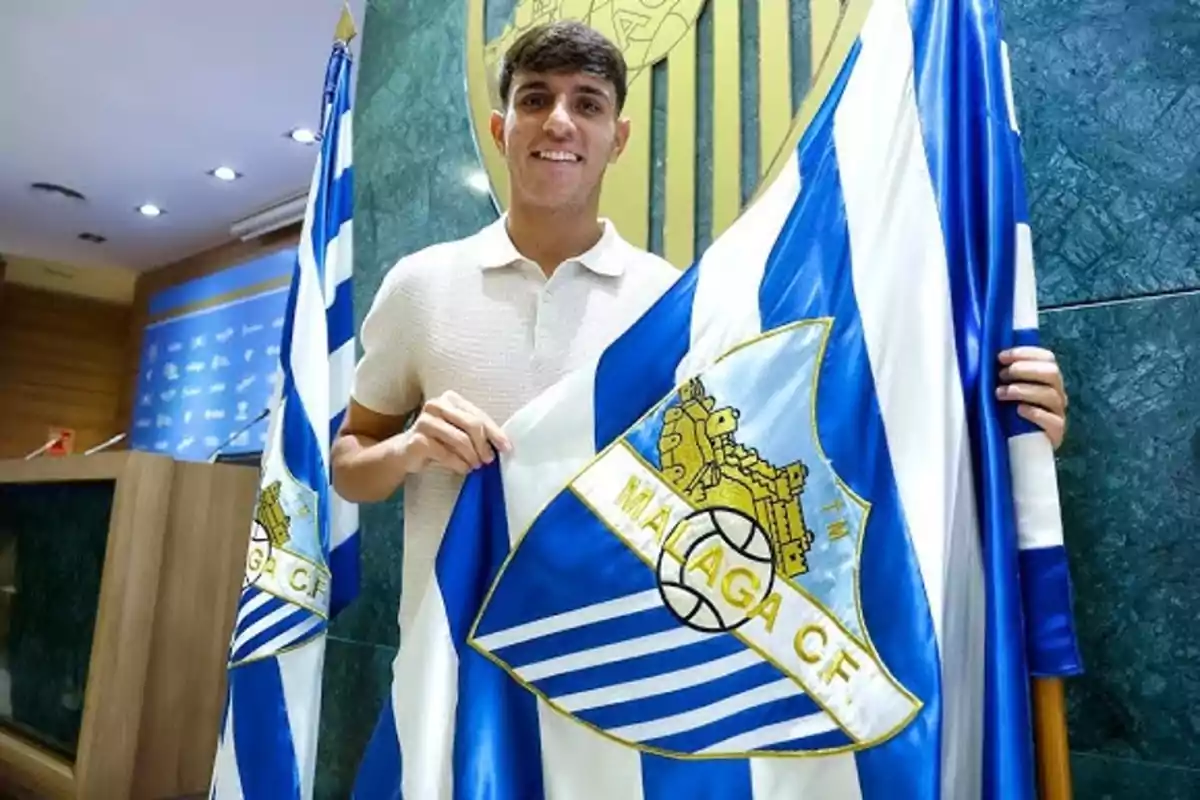 Joven sonriente sostiene una bandera del Málaga CF en una sala de conferencias Joven sonriente sostiene una bandera del Málaga CF en una sala de conferencias