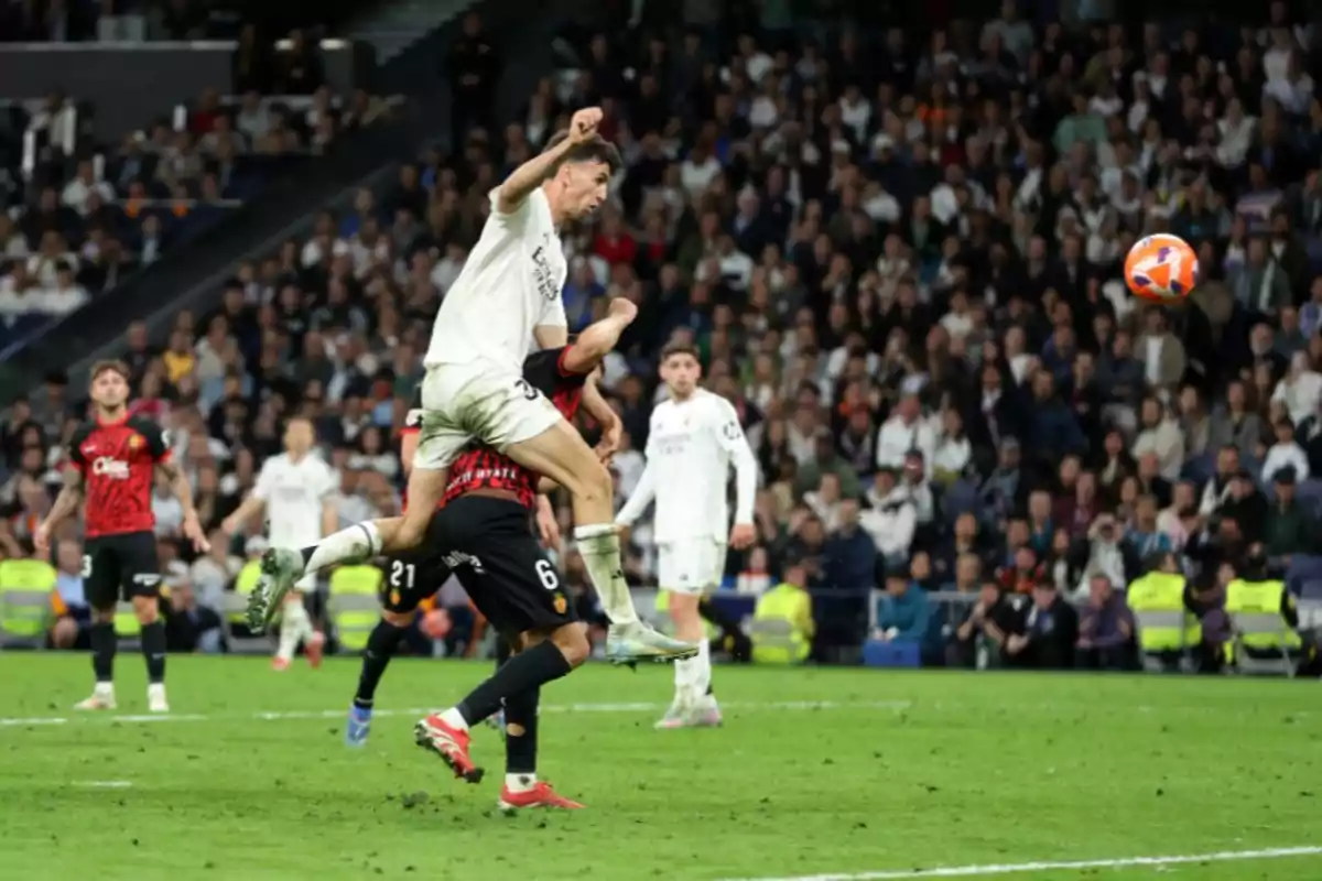 Un jugador de fútbol con uniforme blanco salta para cabecear el balón mientras un jugador del equipo contrario con uniforme rojo y negro intenta defender en un estadio lleno de espectadores. Un jugador de fútbol con uniforme blanco salta para cabecear el balón mientras un jugador del equipo contrario con uniforme rojo y negro intenta defender en un estadio lleno de espectadores.