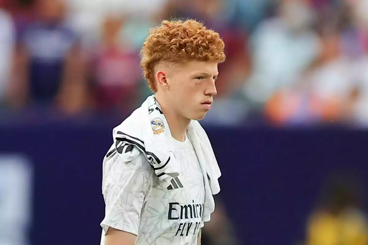 Un joven con cabello rizado y rojizo lleva una camiseta blanca con un logotipo y una toalla sobre el hombro en un entorno deportivo. Un joven con cabello rizado y rojizo lleva una camiseta blanca con un logotipo y una toalla sobre el hombro en un entorno deportivo.