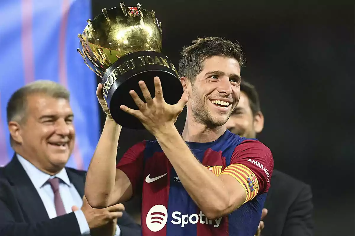 Sergi Roberto sonríe mientras sostiene un trofeo dorado con la inscripción "TROFEU JOAN GAMPER" en el estadio, con personas aplaudiendo en el fondo.