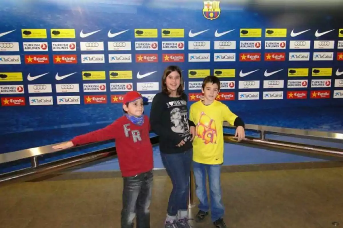 Three children pose in front of a panel with sponsor logos and a soccer team crest. Three children pose in front of a panel with sponsor logos and a soccer team crest.