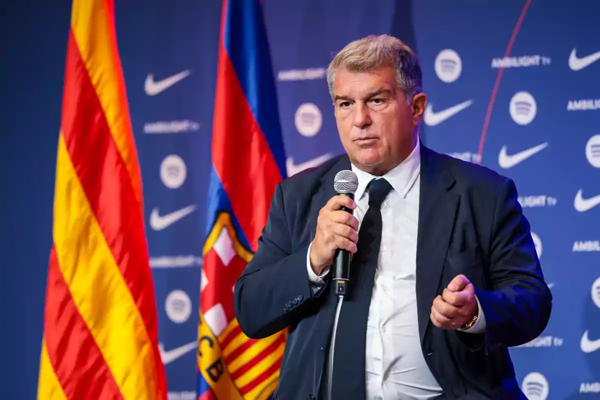 A man in a suit holding a microphone in front of flags and a background with logos.