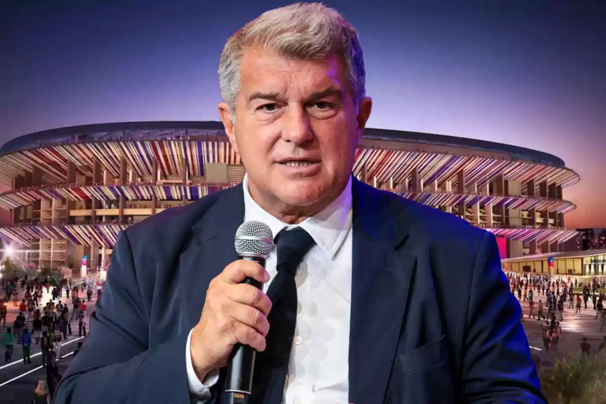 A man in a suit and tie holds a microphone in front of an illuminated stadium at dusk.