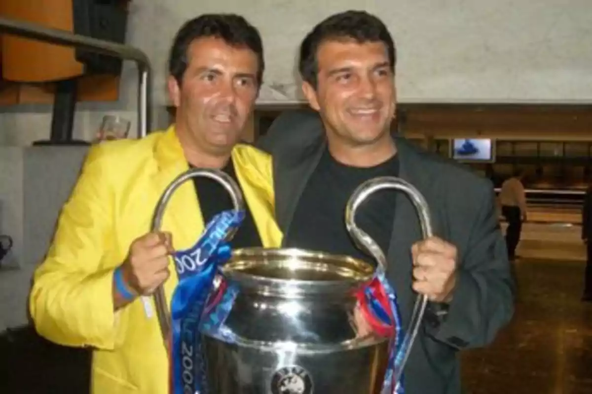 Two people smile as they hold a large silver trophy with handles.