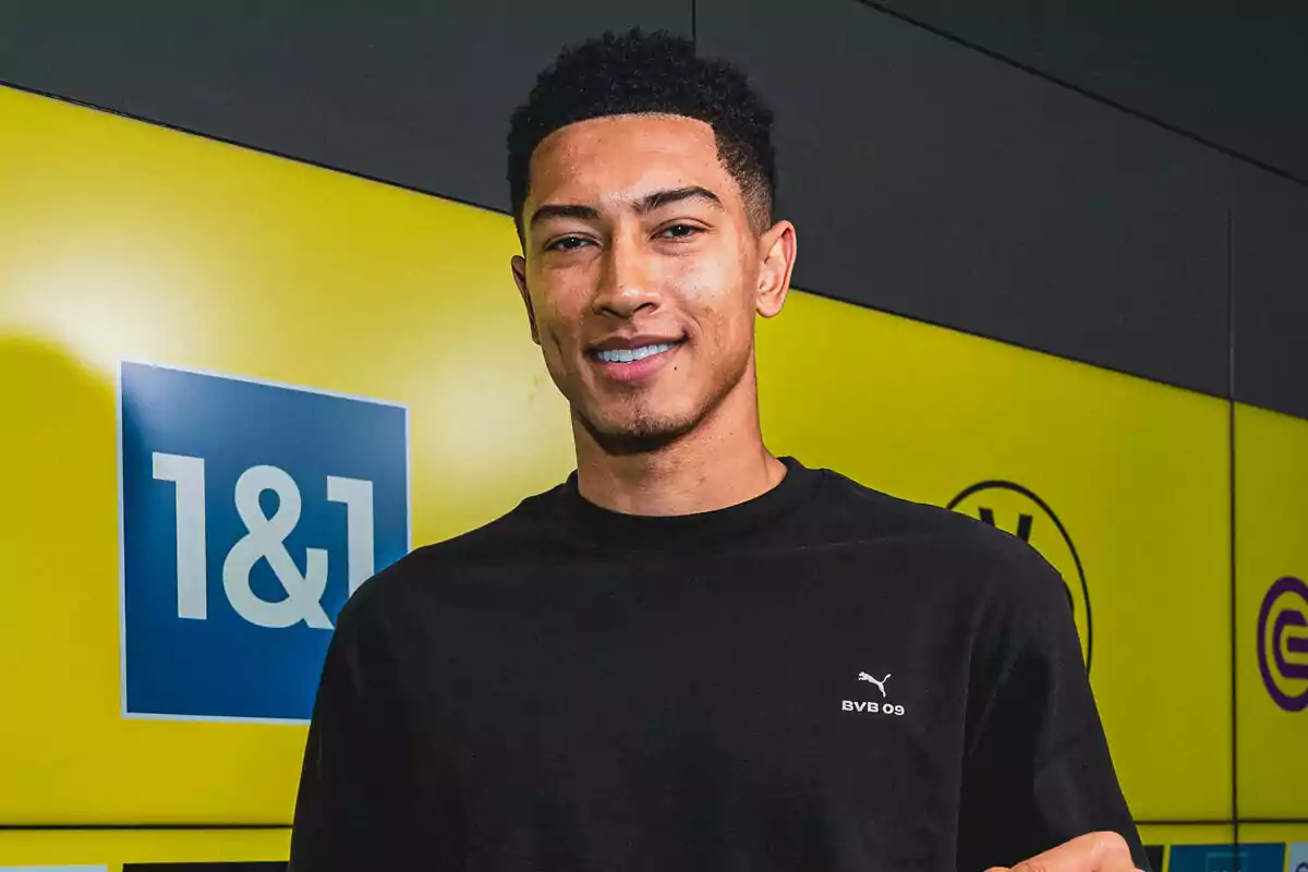 Joven con camiseta negra de Puma sonriendo frente a un fondo amarillo con logotipos. Joven con camiseta negra de Puma sonriendo frente a un fondo amarillo con logotipos.