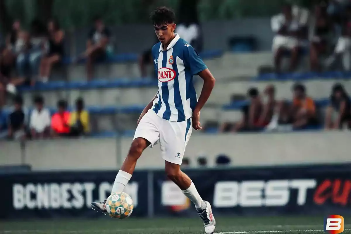 Un jugador de fútbol con uniforme azul y blanco controla el balón en un campo mientras el público observa desde las gradas.