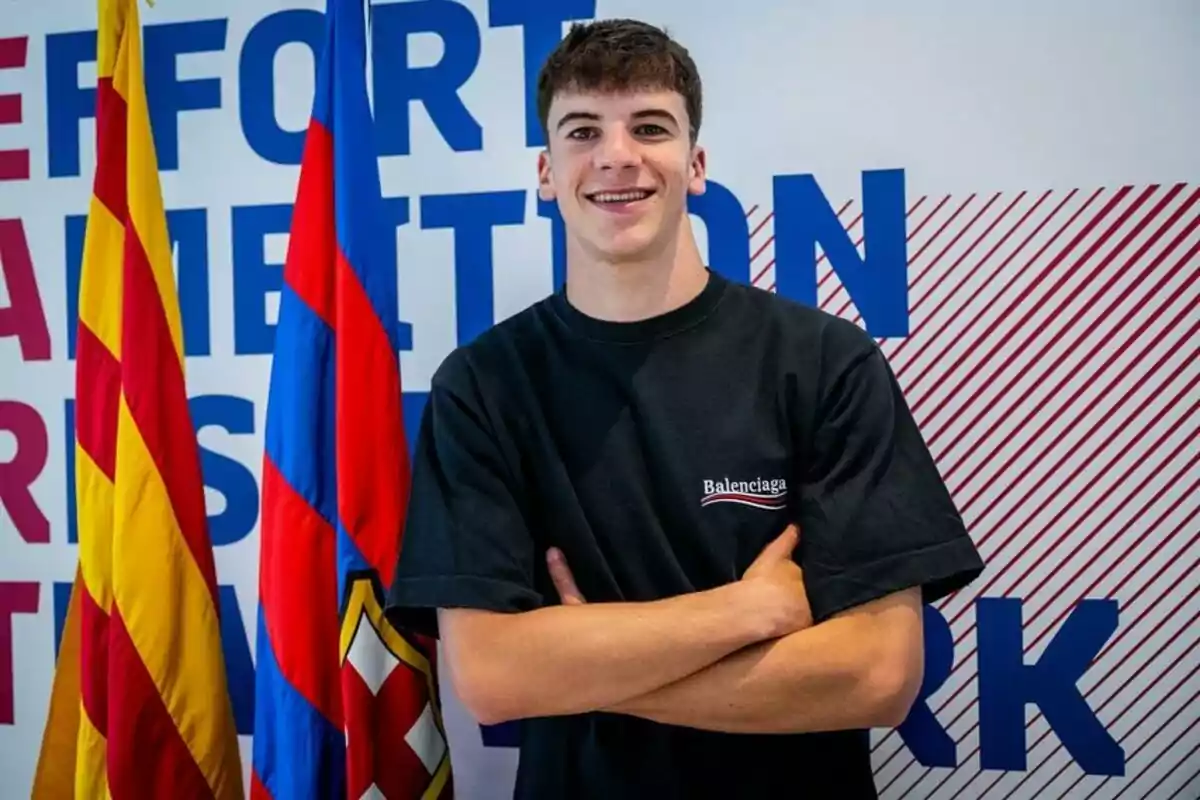 Un joven con camiseta negra de pie frente a banderas y un fondo con texto en azul y rojo. Un joven con camiseta negra de pie frente a banderas y un fondo con texto en azul y rojo.
