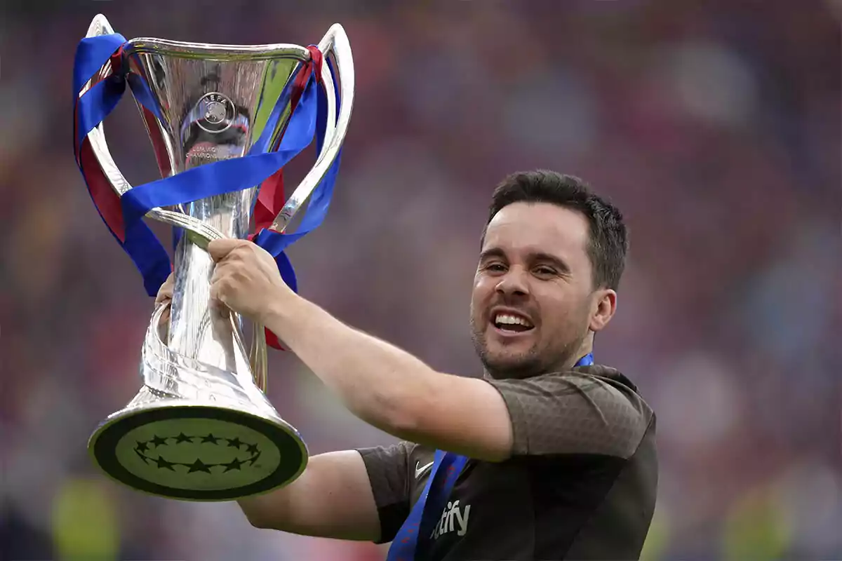 Smiling man lifting a silver trophy adorned with blue and red ribbons in a crowded stadium