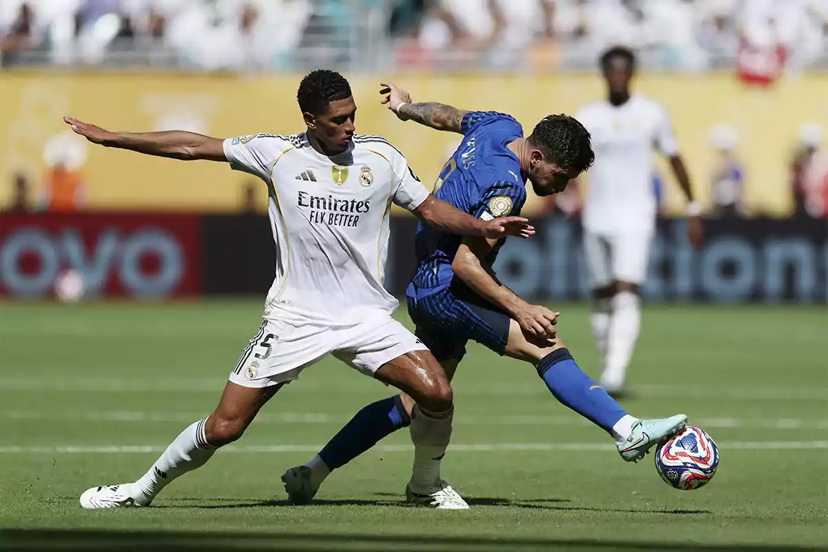 Dos futbolistas disputan el balón durante un partido en un estadio lleno, uno con uniforme blanco y el otro con uniforme azul Dos futbolistas disputan el balón durante un partido en un estadio lleno, uno con uniforme blanco y el otro con uniforme azul