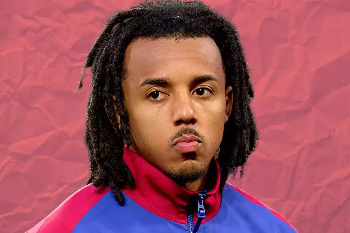 Un hombre con rastas lleva una chaqueta deportiva azul y roja, con un fondo rojo arrugado.