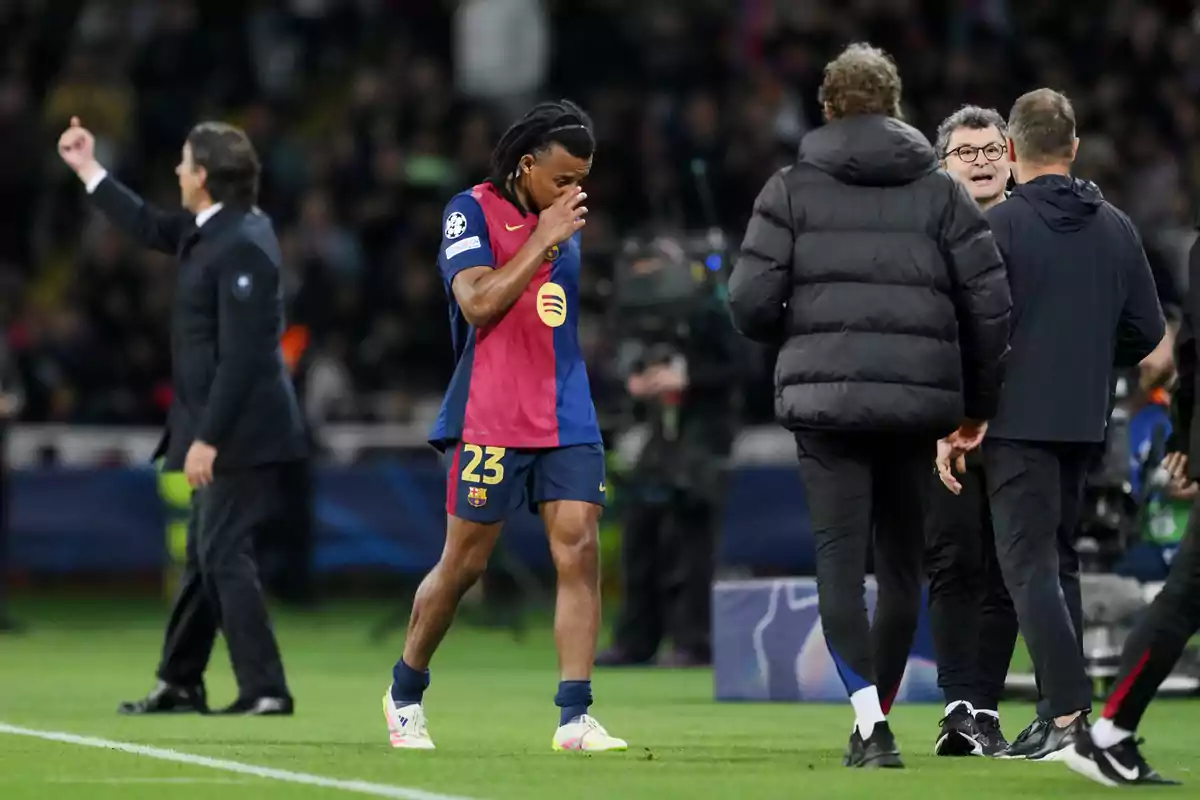 Un jugador de fútbol con el uniforme del Barcelona camina por el campo mientras se toca la cara, rodeado de entrenadores y personal técnico. Un jugador de fútbol con el uniforme del Barcelona camina por el campo mientras se toca la cara, rodeado de entrenadores y personal técnico.