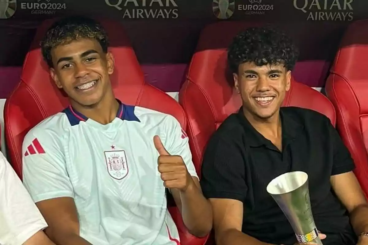 Dos jóvenes sonrientes sentados en sillas rojas, uno lleva una camiseta deportiva blanca y muestra un gesto de aprobación con el pulgar hacia arriba, mientras que el otro sostiene un trofeo y viste una camisa negra. Dos jóvenes sonrientes sentados en sillas rojas, uno lleva una camiseta deportiva blanca y muestra un gesto de aprobación con el pulgar hacia arriba, mientras que el otro sostiene un trofeo y viste una camisa negra.