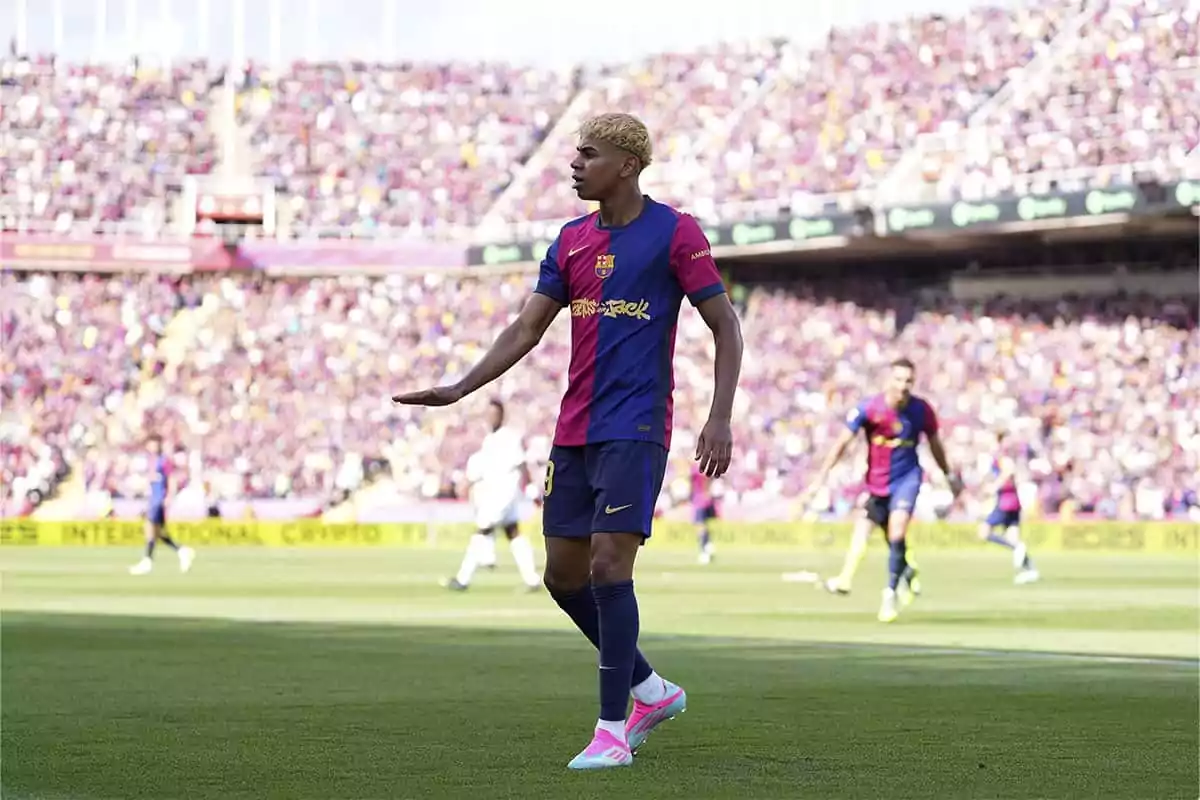 Un jugador de fútbol con uniforme azul y granate está en el campo durante un partido, con el estadio lleno de espectadores al fondo.