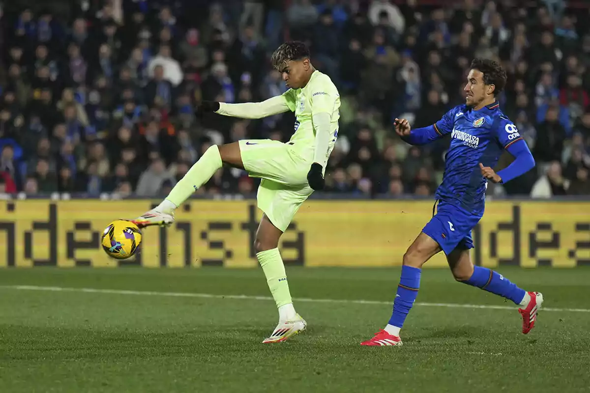 A soccer player in a green uniform controls the ball as an opponent in a blue uniform approaches in a stadium full of spectators.