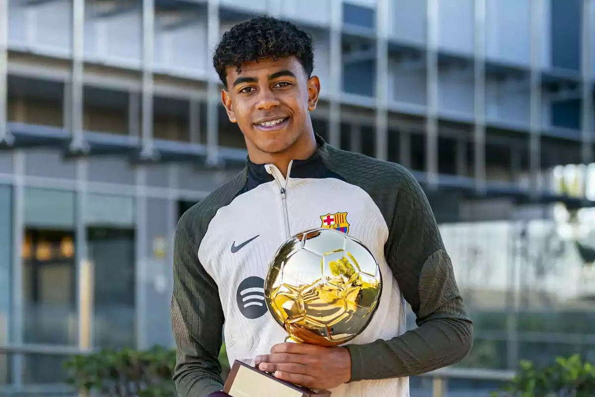 Lamine Yamal con una chaqueta del FC Barcelona sostiene un trofeo dorado frente a un edificio moderno.