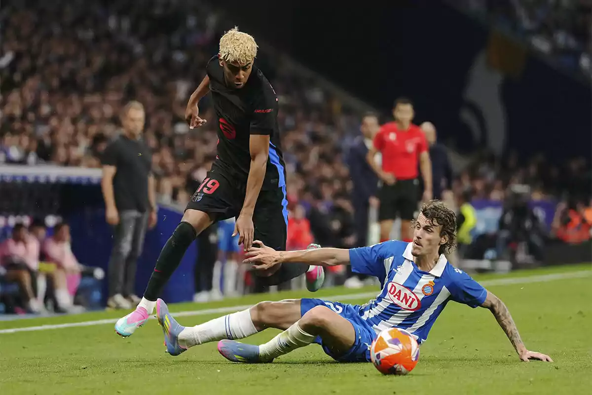Dos jugadores de fútbol compiten por el balón en un partido, uno de ellos está en el suelo intentando interceptar al otro que lleva el balón. Dos jugadores de fútbol compiten por el balón en un partido, uno de ellos está en el suelo intentando interceptar al otro que lleva el balón.