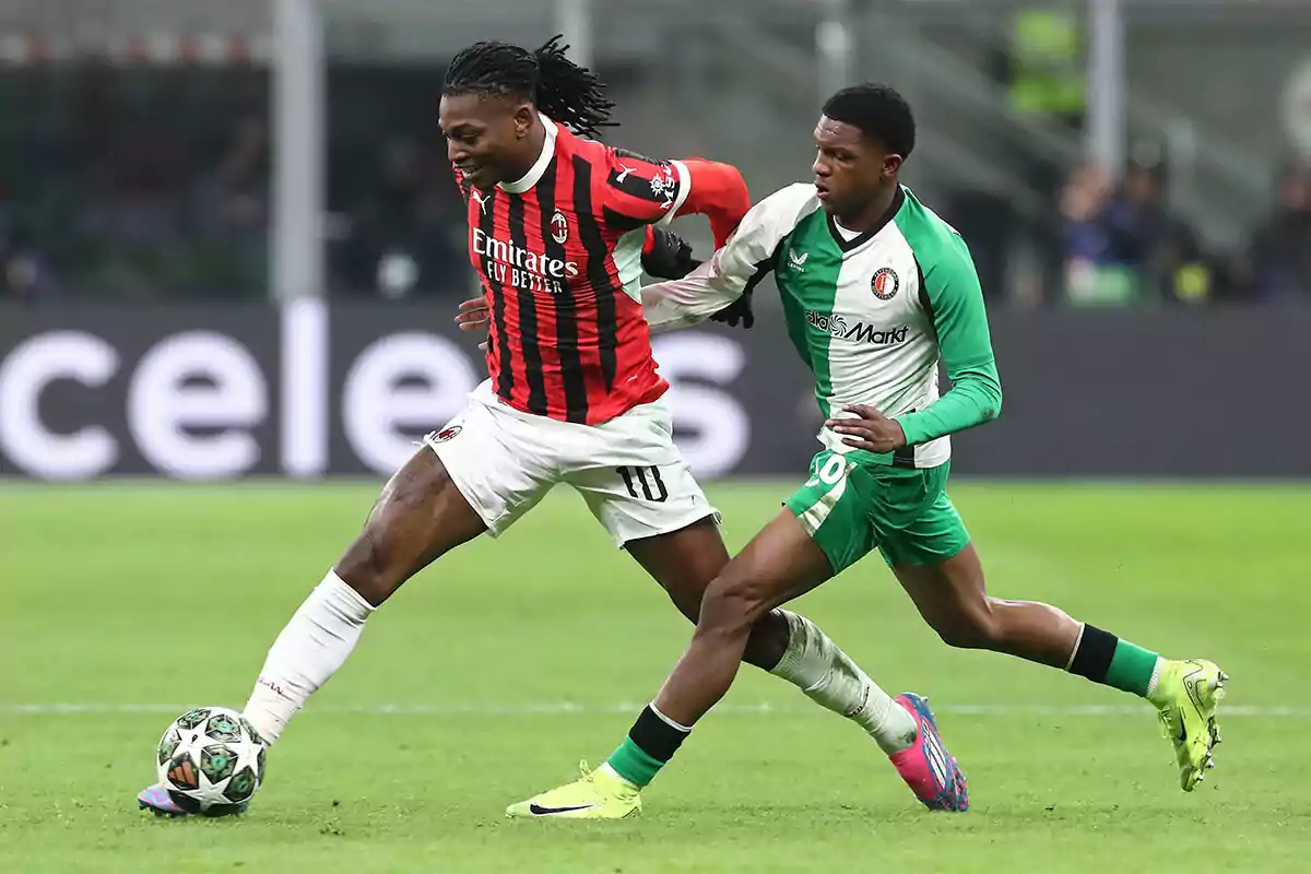 Two players compete for the ball in a match, one in a red and black uniform and the other in a green and white uniform.