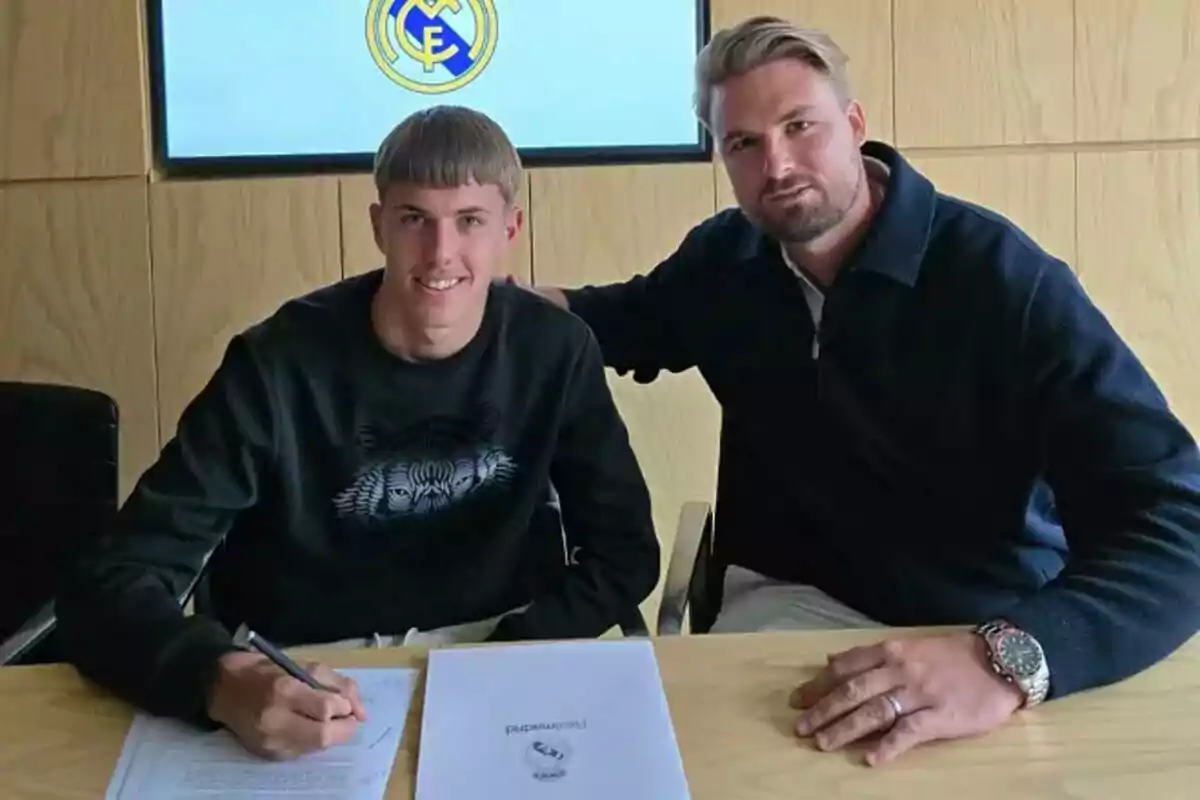 Un joven firmando un documento junto a otra persona en una oficina con el logo del Real Madrid en el fondo. Un joven firmando un documento junto a otra persona en una oficina con el logo del Real Madrid en el fondo.