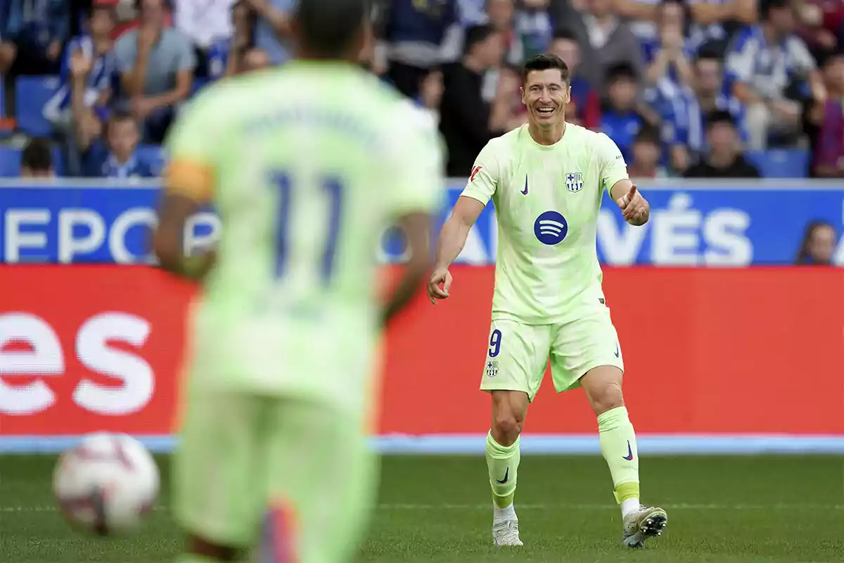 Lewandowski con uniforme verde claro sonríe mientras se dirige hacia Raphinha en el campo durante un partido.