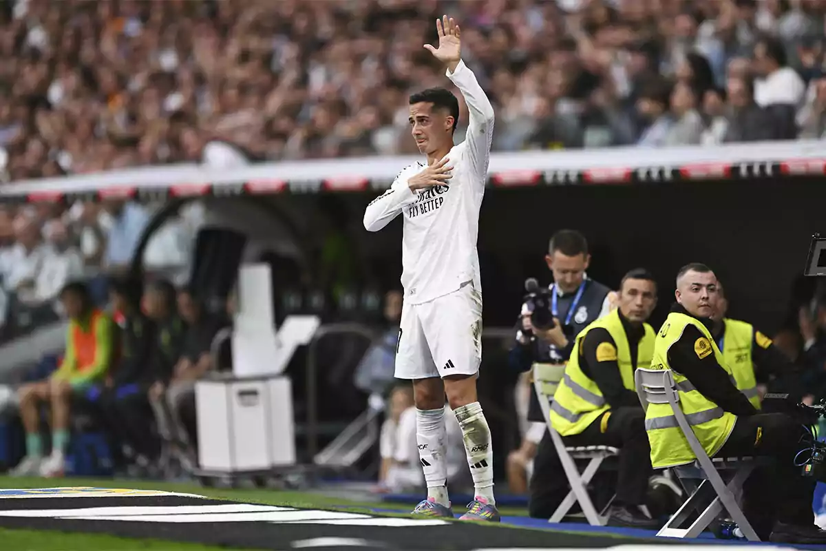 A player in a white uniform waves to the crowd from the field while several people, including photographers and security personnel, watch from the sidelines. A player in a white uniform waves to the crowd from the field while several people, including photographers and security personnel, watch from the sidelines.
