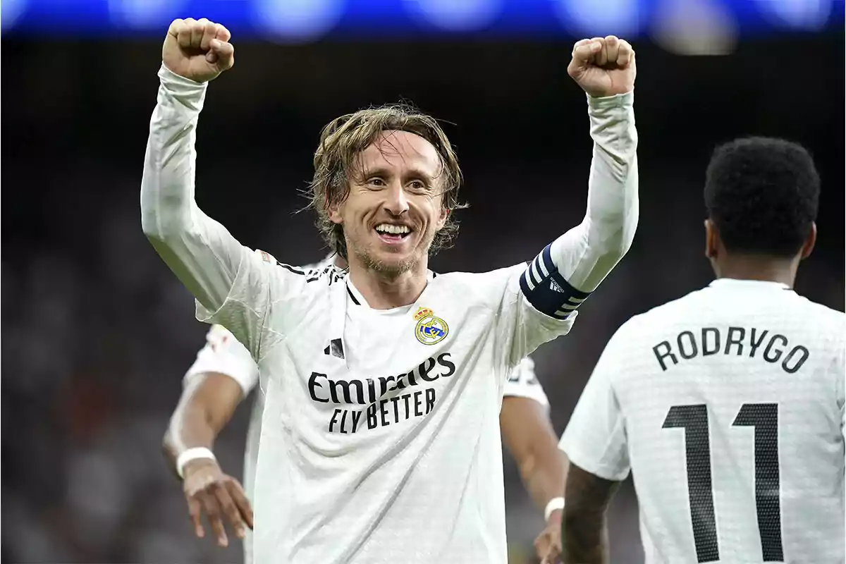 Un jugador de fútbol con uniforme blanco celebra con los brazos en alto mientras otro jugador está de espaldas.