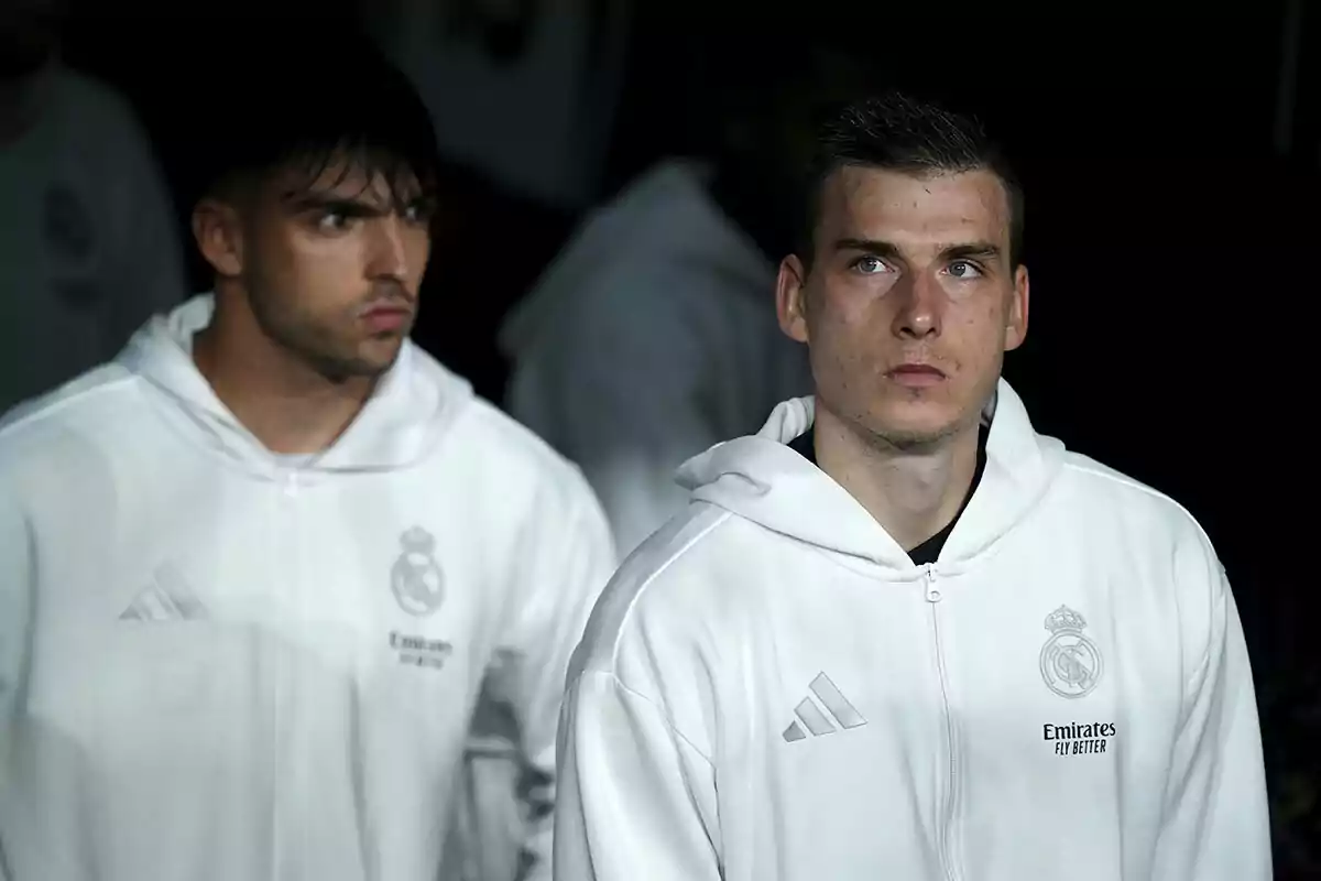 Dos personas con sudaderas blancas del Real Madrid miran hacia adelante con expresiones serias. Dos personas con sudaderas blancas del Real Madrid miran hacia adelante con expresiones serias.
