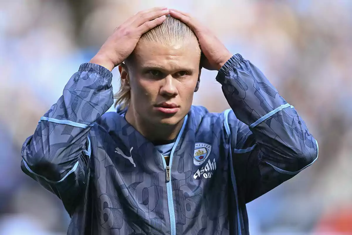 Erling Haaland con una chaqueta azul del Manchester City se sostiene la cabeza con ambas manos en un campo de juego.