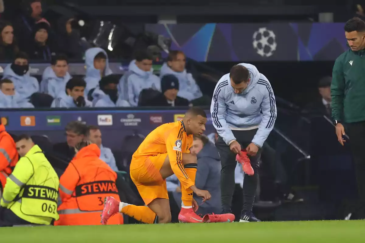 A player in an orange uniform gets ready to enter the field while an assistant helps him with his cleats, with the bench and security personnel visible in the background.