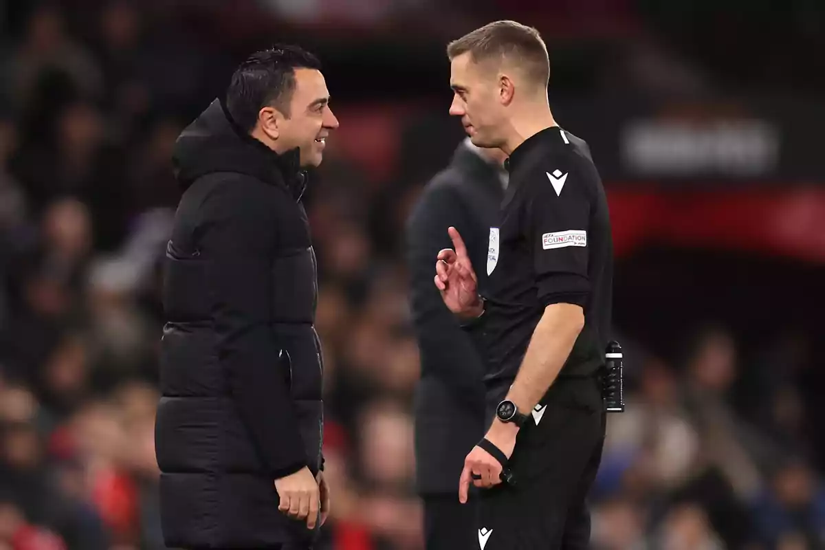 A man in a black jacket talks with a referee dressed in black on a soccer field. A man in a black jacket talks with a referee dressed in black on a soccer field.