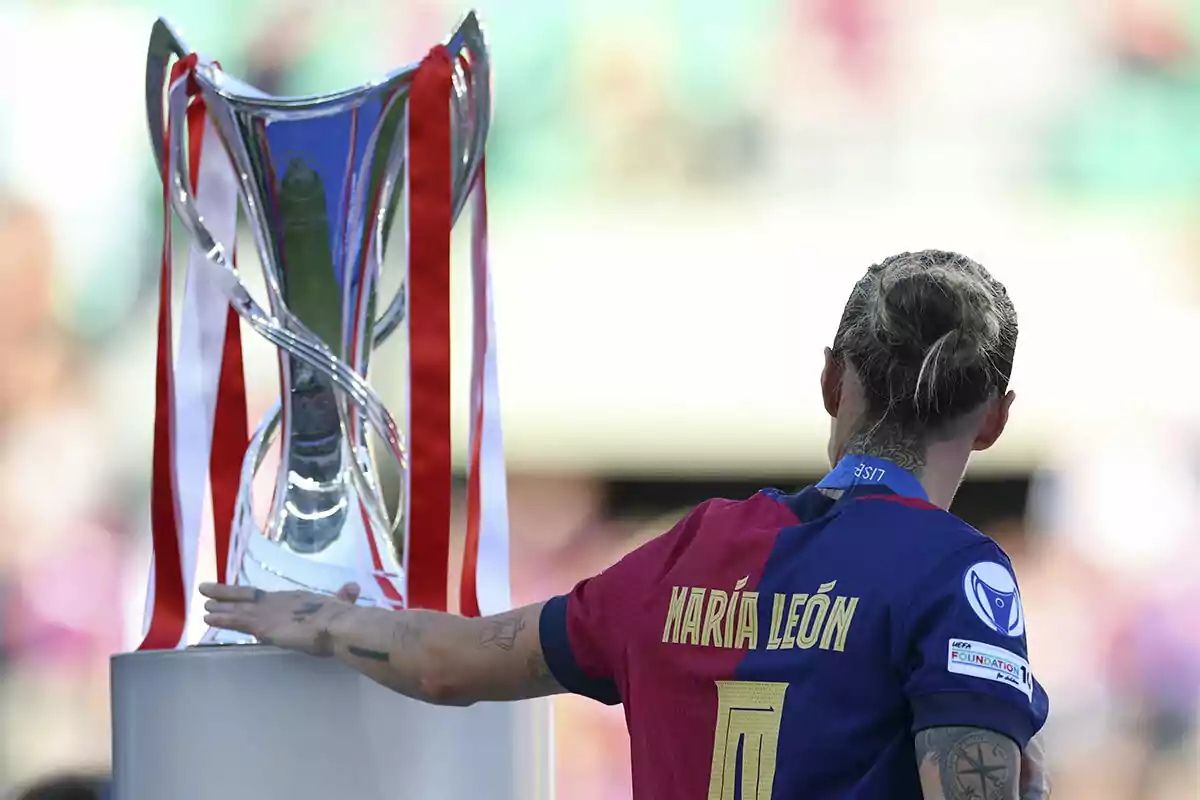 Futbolista con el nombre María León en la camiseta toca un trofeo grande decorado con cintas rojas y blancas Futbolista con el nombre María León en la camiseta toca un trofeo grande decorado con cintas rojas y blancas