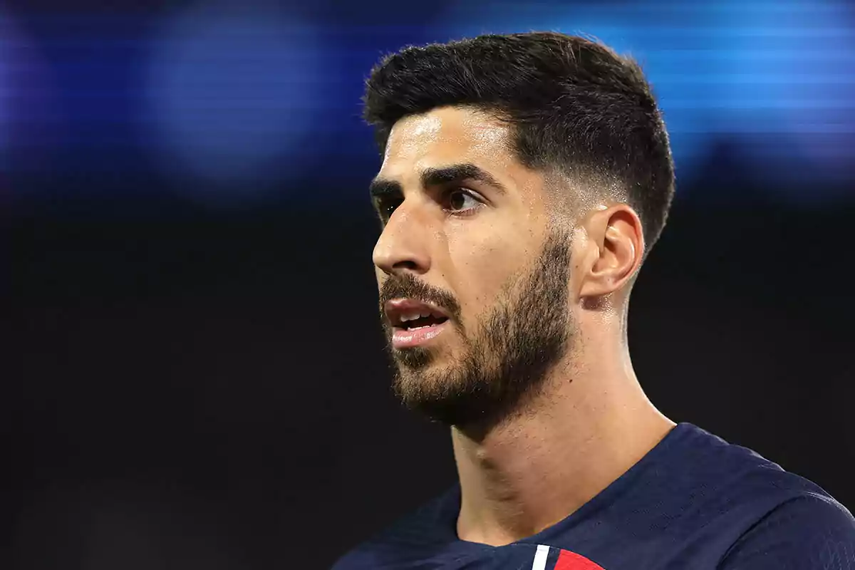 Marco Asensio with a beard and short hair looks forward in a floodlit stadium. Marco Asensio with a beard and short hair looks forward in a floodlit stadium.