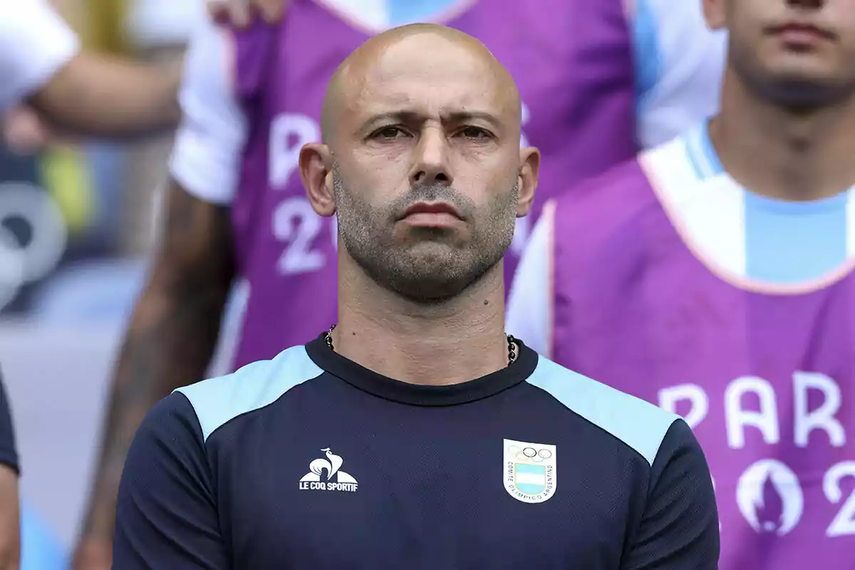 A bald man with a short beard wears a blue and black sports jersey with logos while standing in front of a background with people and purple colors.