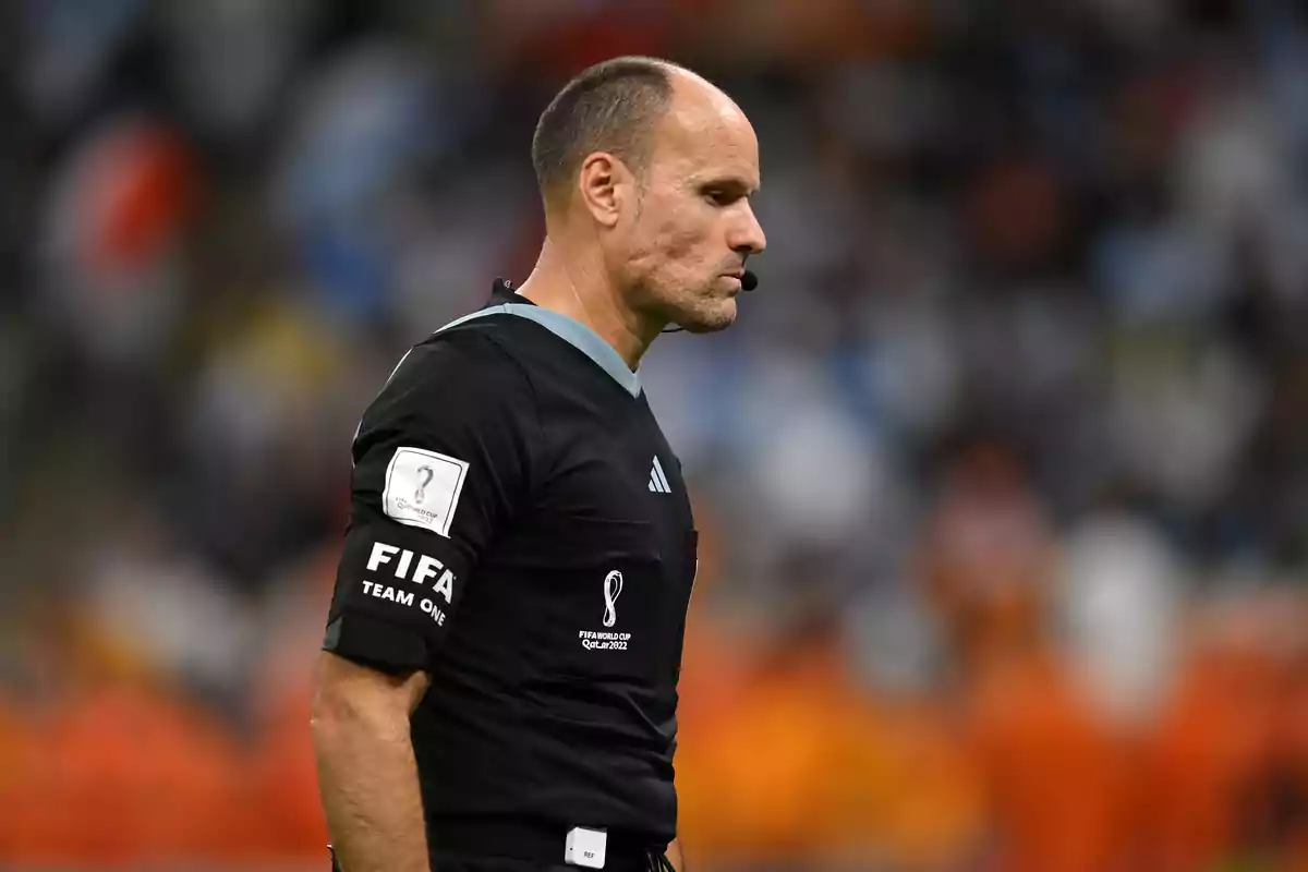 A soccer referee on the field during a match wearing a black uniform and a microphone in the ear.