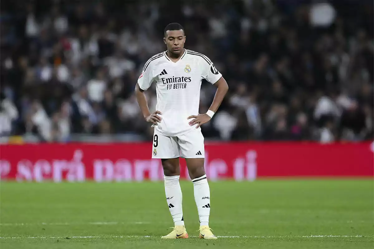 Mbappé en el campo con uniforme blanco y fondo de estadio.