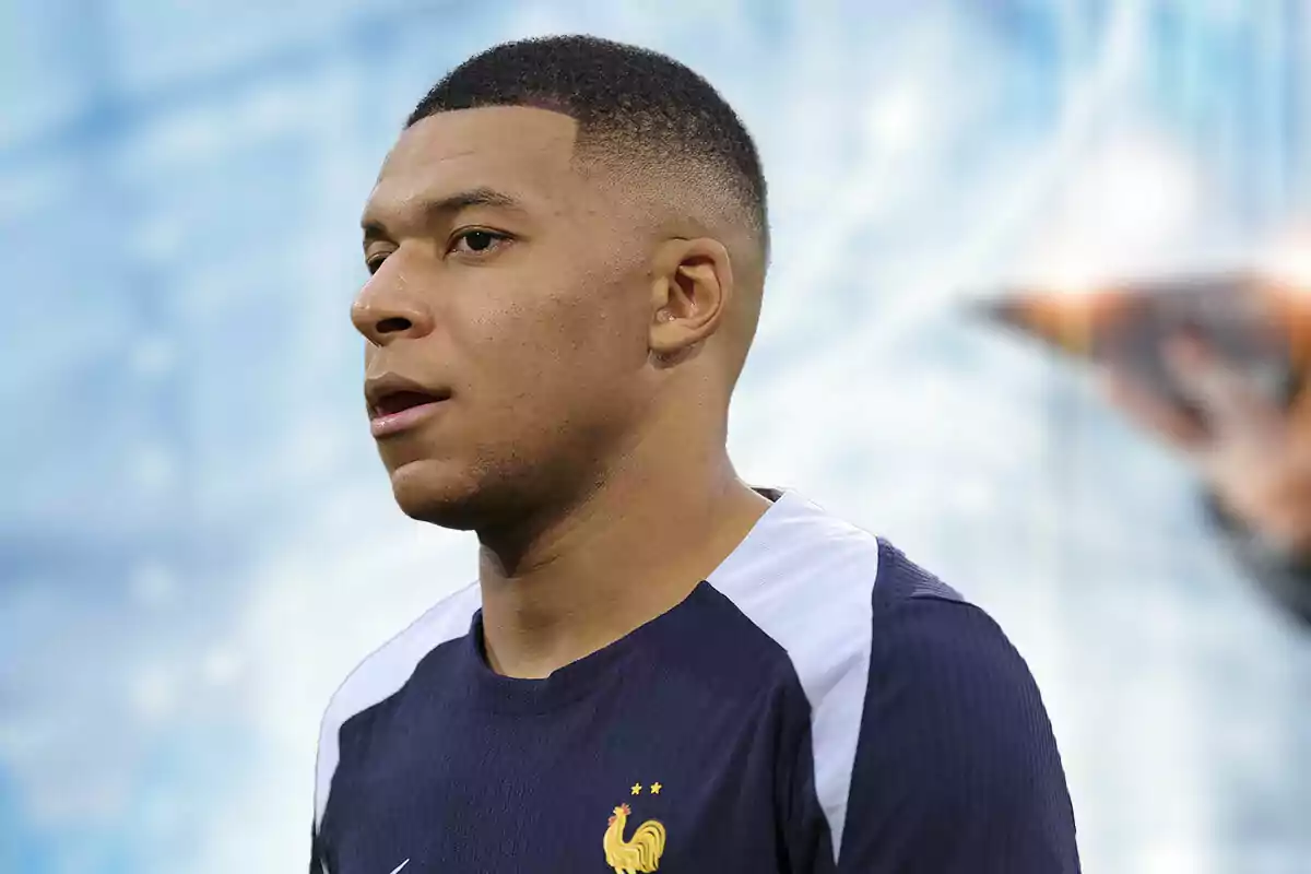 Kylian Mbappé con uniforme azul marino y el escudo de Francia en el pecho, de perfil, con un fondo desenfocado.