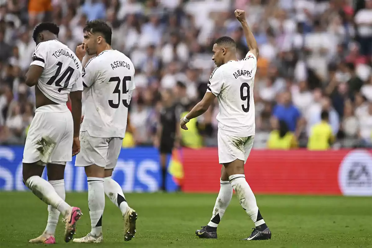 Un grupo de futbolistas en el campo, uno de ellos levanta el brazo en señal de celebración mientras los otros dos caminan juntos. Un grupo de futbolistas en el campo, uno de ellos levanta el brazo en señal de celebración mientras los otros dos caminan juntos.