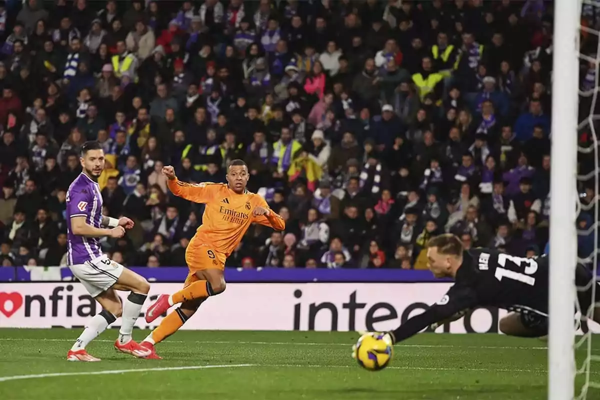A player in an orange uniform attempts to score as the goalkeeper dives to stop the ball in a stadium full of spectators. A player in an orange uniform attempts to score as the goalkeeper dives to stop the ball in a stadium full of spectators.