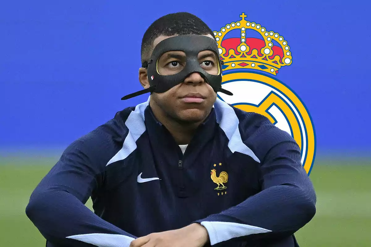 Mbappé con máscara negra sentado frente al logo del Real Madrid.
