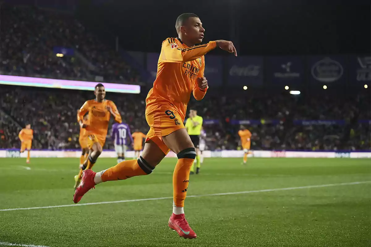 Un jugador de fútbol celebrando un gol en el campo mientras corre con los brazos extendidos, vistiendo un uniforme naranja.