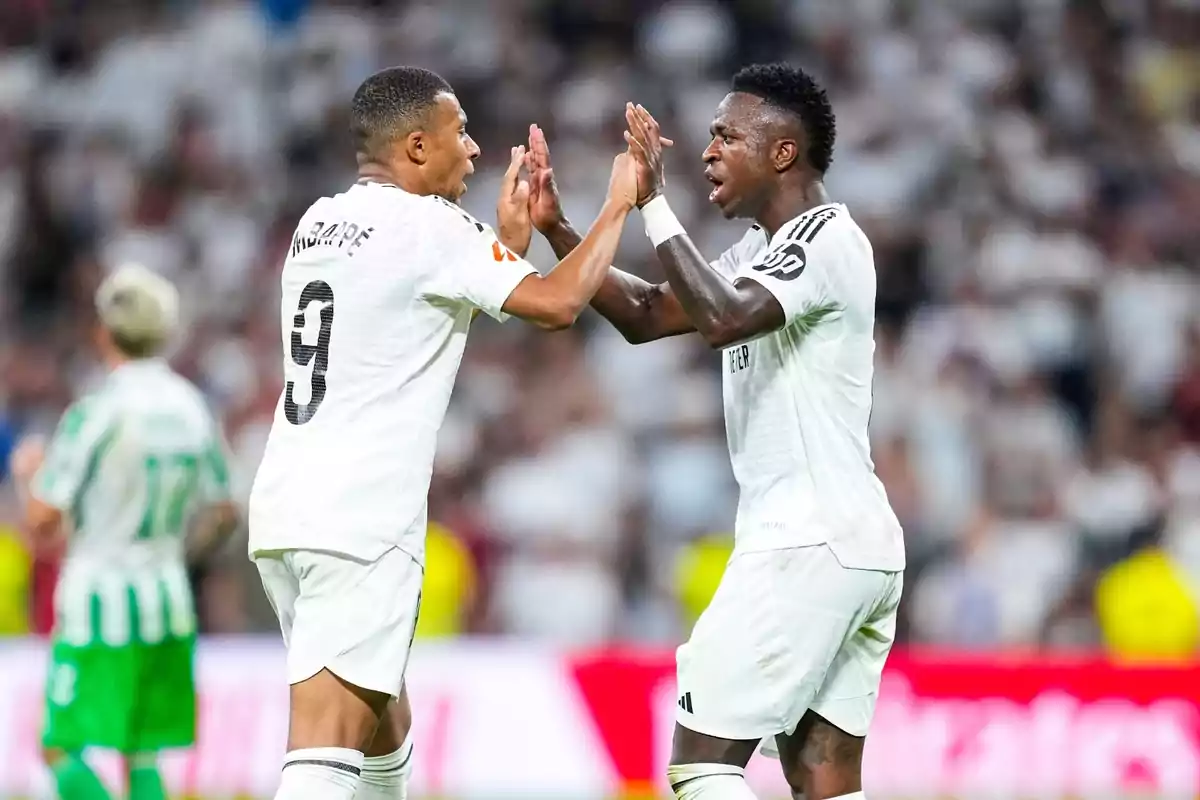 Dos jugadores de fútbol con uniformes blancos celebran en el campo durante un partido.