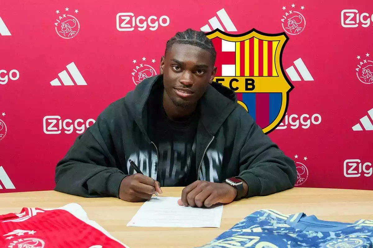 Jorthy Mokio firmando un documento con una camiseta del Ajax, detrás tiene el escudo del FC Barcelona.