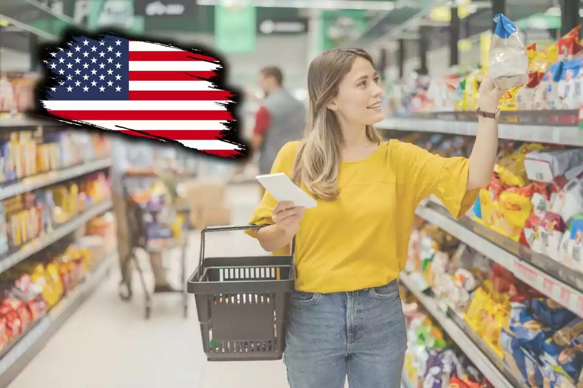 Una mujer con una blusa amarilla sostiene una canasta de compras y un teléfono mientras elige productos en un supermercado; en la esquina superior izquierda hay una bandera de Estados Unidos. Una mujer con una blusa amarilla sostiene una canasta de compras y un teléfono mientras elige productos en un supermercado; en la esquina superior izquierda hay una bandera de Estados Unidos.