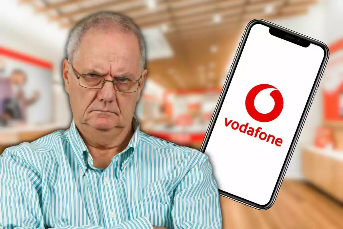 Un hombre mayor con expresión seria y brazos cruzados, con un teléfono móvil que muestra el logo de Vodafone en el fondo de una tienda.