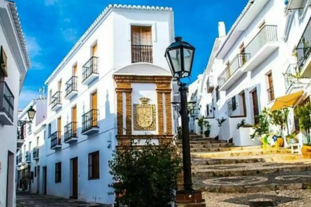 Calle empedrada con casas blancas y balcones en un pueblo andaluz, con un farol y un escudo en la pared.