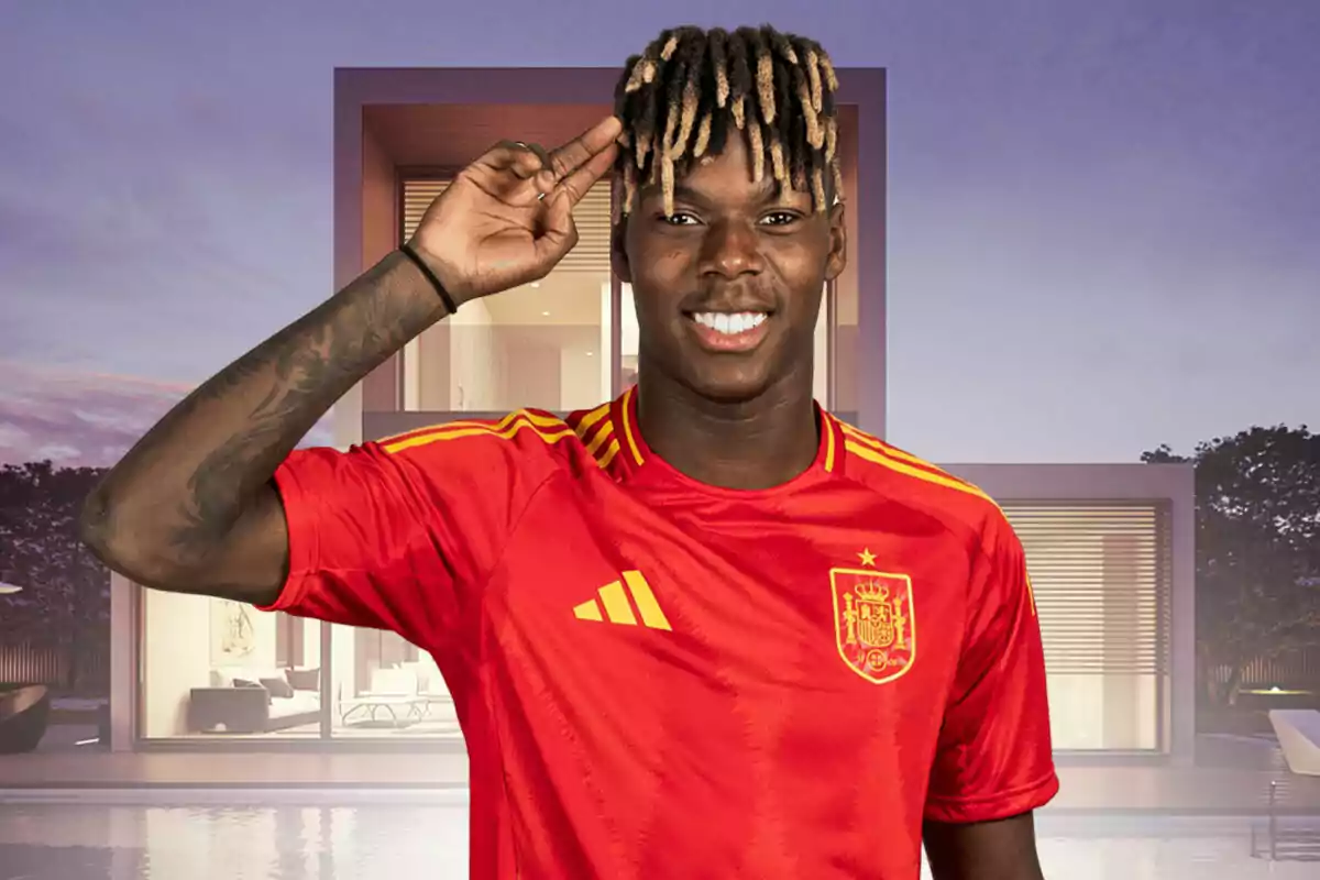 Nico Williams con una camiseta roja de la selección de fútbol de España, sonriendo y haciendo un gesto con la mano en la frente, con una casa moderna de fondo.