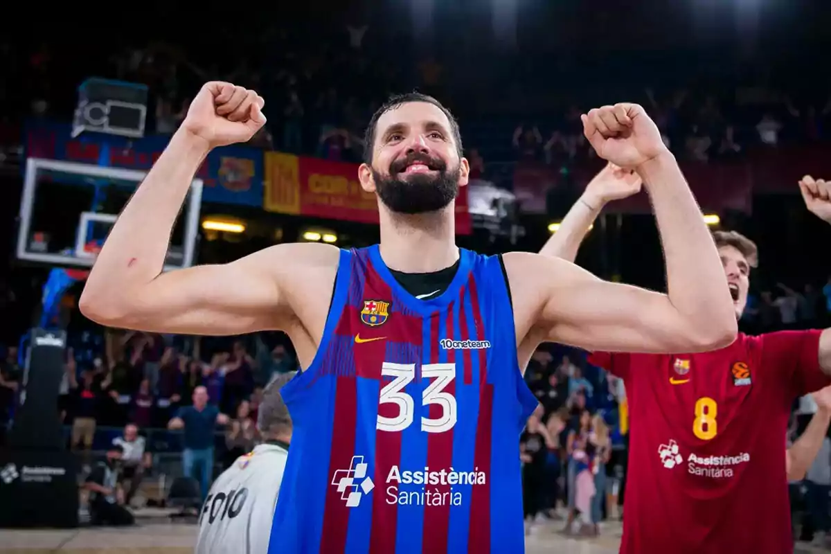 Un jugador de baloncesto con la camiseta del FC Barcelona levanta los brazos en señal de victoria mientras sonríe, rodeado de compañeros y aficionados celebrando en el fondo. Un jugador de baloncesto con la camiseta del FC Barcelona levanta los brazos en señal de victoria mientras sonríe, rodeado de compañeros y aficionados celebrando en el fondo.