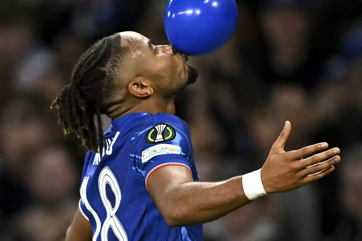 Nkunku con el uniforme azul sostiene un balón azul con la boca durante un partido.