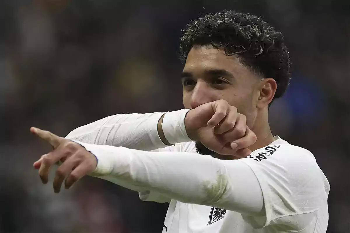 Omar Marmoush in a white uniform celebrates by pointing his finger while covering his mouth with his arm. Omar Marmoush in a white uniform celebrates by pointing his finger while covering his mouth with his arm.